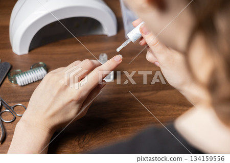A woman performing a DIY manicure by herself, meticulously applying a clear nail polish base coat to her fingernail, using a UV lamp for a perfect finish 134159556