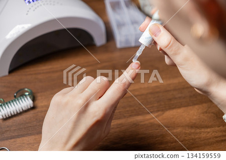 A woman's hands apply clear gel base coat to her bare fingernail, performing a personal, professional-grade self-manicure at home using a UV lamp 134159559