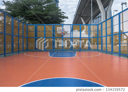 An outdoor basketball court featuring an orange playing surface with blue boundary lines and a basketball hoop. 134159572