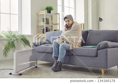 Man sitting at home wrapped in blanket talking by mobile phone warming up with electric heater. Man sitting at home wrapped in blanket talking by mobile phone warming up with electric heater. 134160178