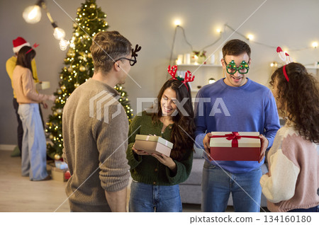 Friends exchanging Christmas gifts at home, laughing together by a festive tree 134160180