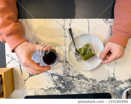 Hands with tea and pistachio baklava from top view. Cozy cafe break, sweet dessert moment, warmth, calm dining atmosphere and gentle pause. 134160498