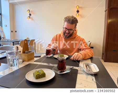 Smiling man with baklava and tea on the table. Dining joy, relaxed break, warm cafe atmosphere, sweetness, communication and comfort. 134160502