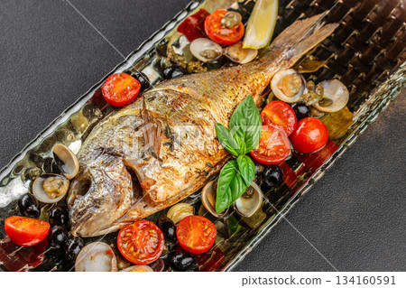 Grilled dorado fish served with cherry tomatoes, olives, clams, capers and fresh basil on a rustic platter, gourmet Mediterranean seafood dish 134160591