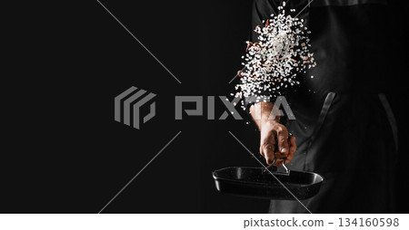 Chef sprinkling coarse salt into a hot pan on dark background with dramatic motion freeze and copy space for professional culinary advertising 134160598