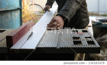 A worker in dark clothes operates a circular saw and cuts a long thin plank into two pieces. A carpenter works in a workshop at a sawing machine during the processing of wood material. A worker in dark clothes operates a circular saw and cuts a long thin plank into two pieces. A carpenter works in a workshop at a sawing machine during the processing of wood material. 134161479