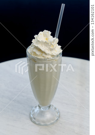 Vanilla milkshake with whipped cream in a tall glass on a white marble table. Vanilla milkshake with whipped cream in a tall glass on a white marble table. 134162101