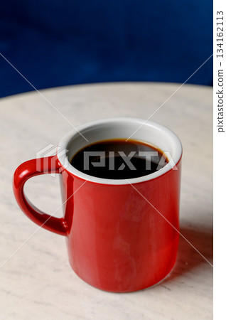 A red ceramic mug with black coffee stands on a white marble table A red ceramic mug with black coffee stands on a white marble table 134162113