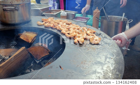 Shrimp sizzling on flat griddle with vendor flipping prawns using spatula, bubbling oil and aromatic spices, outdoor market stall with pots and condiments in background and casual crowd nearby Shrimp sizzling on flat griddle with vendor flipping prawns using spatula, bubbling oil and aromatic spices, outdoor market stall with pots and condiments in background and casual crowd nearby 134162408