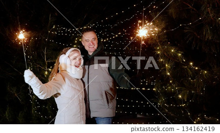 Celebrating winter holiday, couple standing outdoors at night, holding sparklers with glowing fir trees behind, sharing joyful moment during festive season Celebrating winter holiday, couple standing outdoors at night, holding sparklers with glowing fir trees behind, sharing joyful moment during festive season 134162419