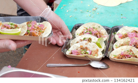 Tightly packed line of foil trays with ready tacos surrounded by utensils and condiments, Busy event food stand where staff prepare and arrange multiple taco portions for eager festival visitors Tightly packed line of foil trays with ready tacos surrounded by utensils and condiments, Busy event food stand where staff prepare and arrange multiple taco portions for eager festival visitors 134162421