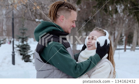 Couple sharing playful moment, dreadlocked man adjusting earmuffs for girlfriend during snowy winter park walk, both laughing 134162432