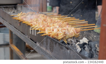 Cooking fires with smoky festival atmosphere, Grilling chicken on skewers amid lively festival heat and smoke, Rows of skewers roasting over embers during animated festival preparations with steady Cooking fires with smoky festival atmosphere, Grilling chicken on skewers amid lively festival heat and smoke, Rows of skewers roasting over embers during animated festival preparations with steady 134162440