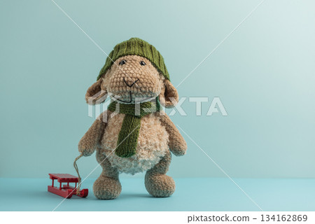 Plush Sheep in Green Hat and Scarf Pulling Red Sled on Blue Background 134162869