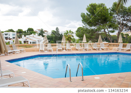 Blue swimming pool with many sun loungers and umbrellas around. Rest zone surrounded by green trees and villas on Mallorca island. Blue swimming pool with many sun loungers and umbrellas around. Rest zone surrounded by green trees and villas on Mallorca island. 134163234