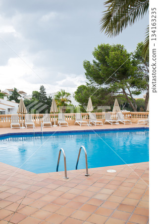 Blue swimming pool with many sun loungers and umbrellas around. Rest zone surrounded by green trees and villas on Mallorca island. Blue swimming pool with many sun loungers and umbrellas around. Rest zone surrounded by green trees and villas on Mallorca island. 134163235