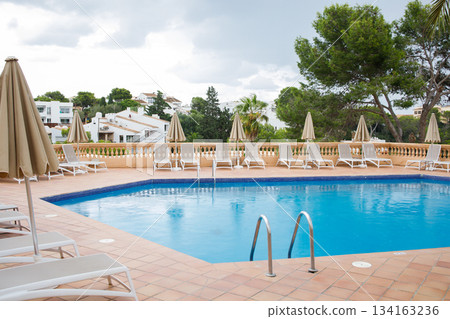 Blue swimming pool with many sun loungers and umbrellas around. Rest zone surrounded by green trees and villas on Mallorca island. Blue swimming pool with many sun loungers and umbrellas around. Rest zone surrounded by green trees and villas on Mallorca island. 134163236