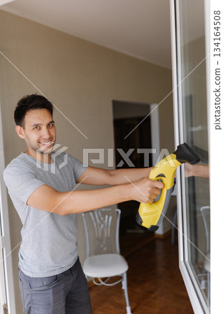 Portrait of friendly cleaning service employee wiping glass surface using cordless handheld vacuum, enjoying chore in apartment, looking at camera. Concept of domestic work, housekeeping and household Portrait of friendly cleaning service employee wiping glass surface using cordless handheld vacuum, enjoying chore in apartment, looking at camera. Concept of domestic work, housekeeping and household 134164508