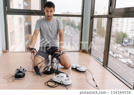 Portrait of frustrated young man crouching among tangled window cleaning robot cables, troubleshooting technical setup near expansive glass windows in living space, puzzled looking down. Portrait of frustrated young man crouching among tangled window cleaning robot cables, troubleshooting technical setup near expansive glass windows in living space, puzzled looking down. 134164556