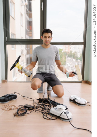 Vertical portrait of young man kneeling on floor, holding cordless window vacuum cleaner and wiper, preparing robot window cleaners and other appliances for cleaning windows, looking at camera. 134164557