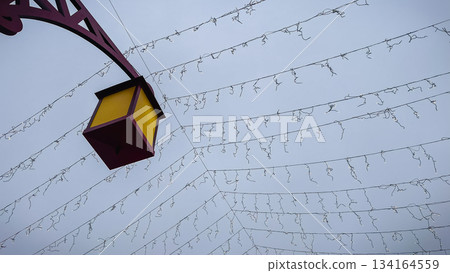 Decorative street lantern against a gray winter sky with a ceiling of suspended holiday light cables and wires Decorative street lantern against a gray winter sky with a ceiling of suspended holiday light cables and wires 134164559