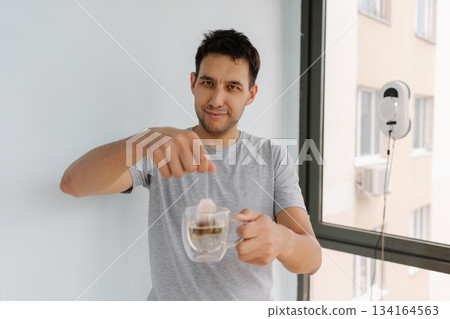 Happy smiling man looking at camera brewing tea near automated window-cleaning robot, enjoying peaceful moment in bright, contemporary living space with sunlight streaming through glass Happy smiling man looking at camera brewing tea near automated window-cleaning robot, enjoying peaceful moment in bright, contemporary living space with sunlight streaming through glass 134164563