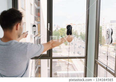 Back view of man controlling cleaning robots on skyscraper window using remote control, showcasing innovative technology for high-rise window cleaning with cityscape view. Concept of cleaning house Back view of man controlling cleaning robots on skyscraper window using remote control, showcasing innovative technology for high-rise window cleaning with cityscape view. Concept of cleaning house 134164565