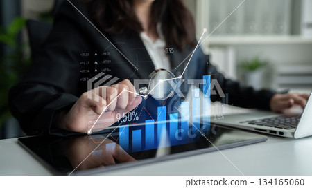 Businesswoman analyzing financial data through a digital tablet with graphics, magnifying glass, and laptop in a modern office setting Scalp 134165060