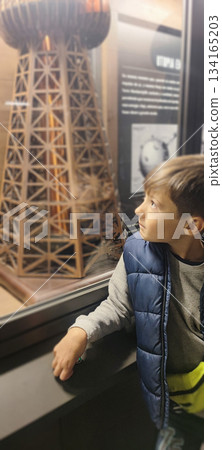 A young boy looks at an information panel and a large model of a structure behind glass at an exhibition and presses a button. The concept is a children's museum experience and scientific research. 134165203