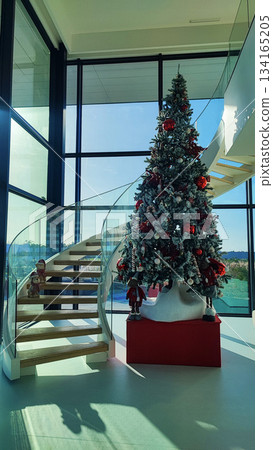 A modern interior with a large decorated Christmas tree next to a curved glass staircase in a bright room. The concept of Christmas in contemporary design, festivity, and a seasonal atmosphere. A modern interior with a large decorated Christmas tree next to a curved glass staircase in a bright room. The concept of Christmas in contemporary design, festivity, and a seasonal atmosphere. 134165205