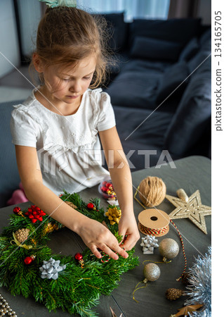 Little girl finishing her handmade Christmas wreath by tying a string to hang it. Final step of a festive DIY project captured at home in a cozy family setting. Little girl finishing her handmade Christmas wreath by tying a string to hang it. Final step of a festive DIY project captured at home in a cozy family setting. 134165705