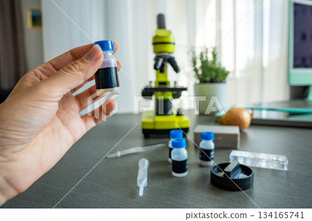 A person holding a small sample vial in front of a yellow microscope, with more scientific supplies on the table. Concept of science education and home experiments for kids 134165741