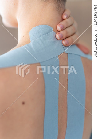 Close up view of young woman with kinesiology tape applied to her neck and shoulder holding her neck with her hand. Physical therapy and muscle recovery through modern rehabilitative care Close up view of young woman with kinesiology tape applied to her neck and shoulder holding her neck with her hand. Physical therapy and muscle recovery through modern rehabilitative care 134165764
