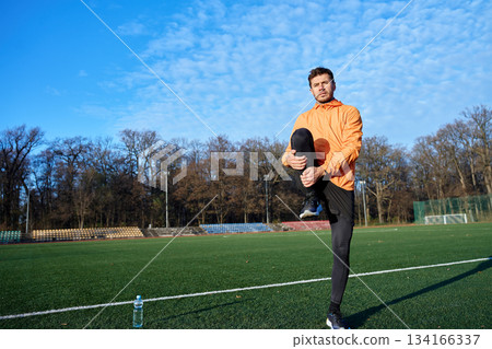 Man stretching leg while standing on sports field at outdoor stadium Man stretching leg while standing on sports field at outdoor stadium 134166337