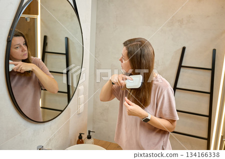 Woman brushing long hair while standing in front of bathroom mirror at home Woman brushing long hair while standing in front of bathroom mirror at home 134166338