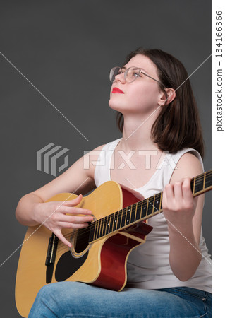 Guitarist with serene countenance plays acoustic guitar during performance, demonstrating profound level of engagement in creative expression. European woman musician, from zoomers, wearing eyeglasses 134166366