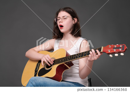 Woman guitarist and singer songwriter plays guitar and singing song with eyes closed, completely surrendering to creative process during rehearsal. Woman guitarist, belonging to Gen Z, wearing glasses Woman guitarist and singer songwriter plays guitar and singing song with eyes closed, completely surrendering to creative process during rehearsal. Woman guitarist, belonging to Gen Z, wearing glasses 134166368