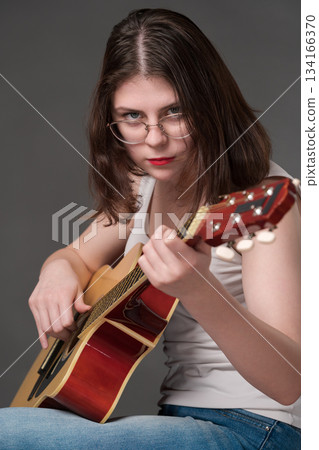 Female rock musician plays classical six string acoustic guitar, performing technically challenging piece of music. Young adult woman guitar player wearing glasses and directing gaze toward camera 134166370