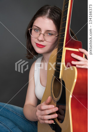 Woman guitarist with positive emotional expression, holding guitar vertically and gently embracing musical instrument. 18 year old woman musician belonging to Gen Z, wearing glasses, white tank top Woman guitarist with positive emotional expression, holding guitar vertically and gently embracing musical instrument. 18 year old woman musician belonging to Gen Z, wearing glasses, white tank top 134166373