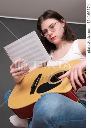 Woman musician holds sheet music in front of her, deciphering and analyzing new musical creation in creative process during rehearsal. Young guitarist singer songwriter with guitar wearing eyeglasses 134166374
