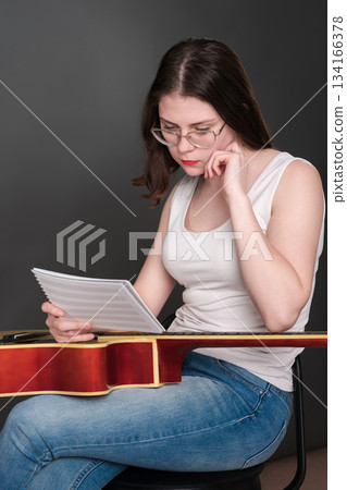 Female guitar player, intrigued by intricacies of her craft, holds musical notes before her, contemplating and interpreting novel musical composition. Singer songwriter in midst of creative process Female guitar player, intrigued by intricacies of her craft, holds musical notes before her, contemplating and interpreting novel musical composition. Singer songwriter in midst of creative process 134166378
