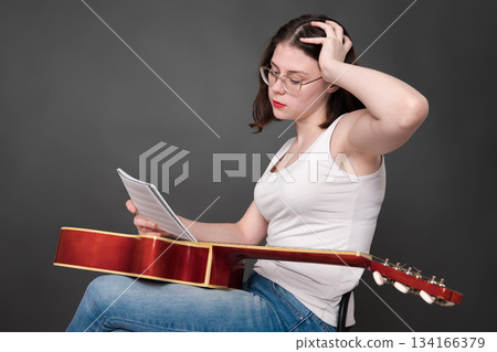 Perplexed female musician holds musical notes in front of her, deciphering and analyzing new musical creation. Female singer songwriter sitting with guitar on lap in creative process during rehearsal 134166379