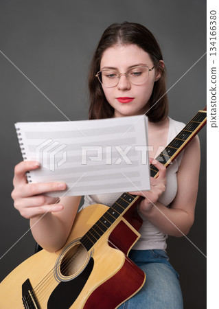 Female guitar player holding lyrics and musical notes in hand. She is positioned in front of body, with eyes focused on reading and studying new musical work. Woman singer songwriter wears eyeglasses 134166380