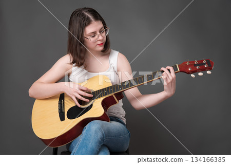 Woman guitarist playing acoustic classical guitar and looking at string. Young adult European woman guitar player, belonging to Generation Z, wearing glasses and white tank top, blue jeans Woman guitarist playing acoustic classical guitar and looking at string. Young adult European woman guitar player, belonging to Generation Z, wearing glasses and white tank top, blue jeans 134166385
