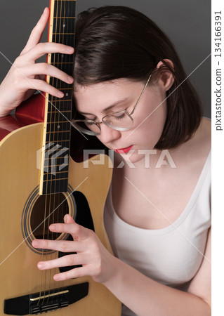 Woman musician with eyes closed, holding guitar vertically next to her and tenderly hugging musical instrument, gently pressing it against body. Female guitarist wearing eyeglasses, white tank top 134166391