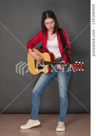 Young woman guitarist playing six string acoustic guitar, standing full length on stage. Young adult female musician with glasses is dressed in red leather jacket, jeans and sneakers 134166409