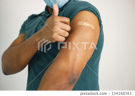 Black man standing isolated on a white background. Man holding a thumb up and showing a band-aid on a shoulder. Man wearing blue shirt and medical face mask. Black man standing isolated on a white background. Man holding a thumb up and showing a band-aid on a shoulder. Man wearing blue shirt and medical face mask. 134168561