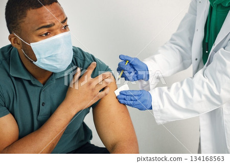 Black man sitting isolated on a white background. Doctor in medical gown holding a syringe. Man wearing blue shirt and medical face mask. 134168563