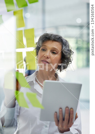 Glass wall, tablet and business woman writing, planning and strategy in office workplace. Sticky notes, brainstorming and senior female professional with technology for working, schedule and info. Glass wall, tablet and business woman writing, planning and strategy in office workplace. Sticky notes, brainstorming and senior female professional with technology for working, schedule and info. 134168564