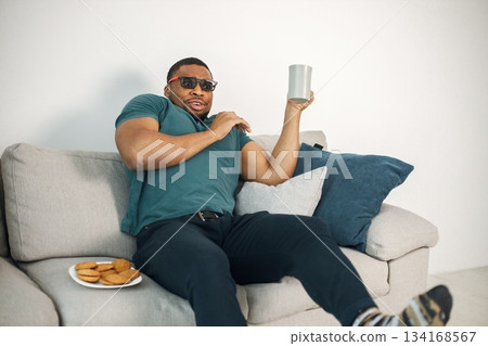 Black man sitting on a coach and holding a cup with a tea. Man wearing blue t-shirt and 3D glasses. Man watching a movie. 134168567